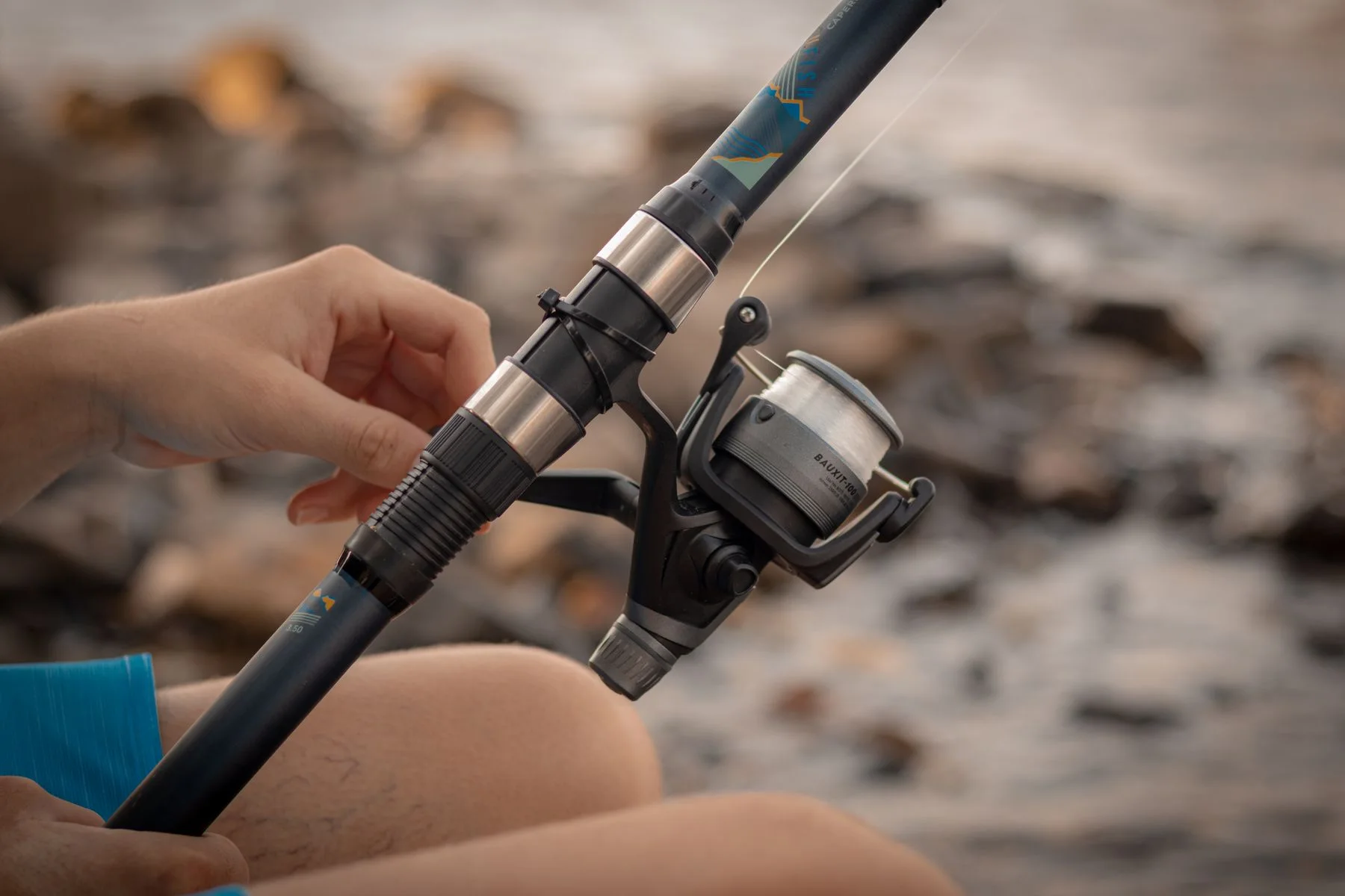 Hands holding a fishing rod and spinning reel beside rocks and water.