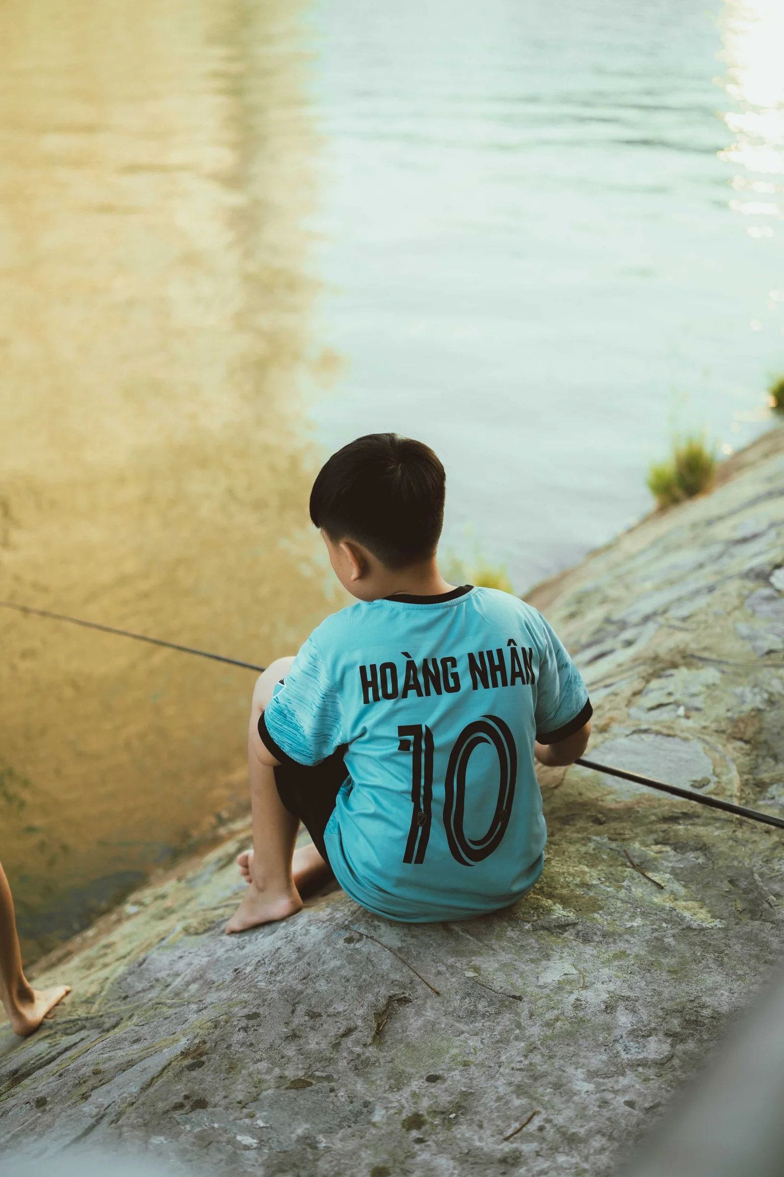 Boy sitting on a riverbank with a fishing rod.