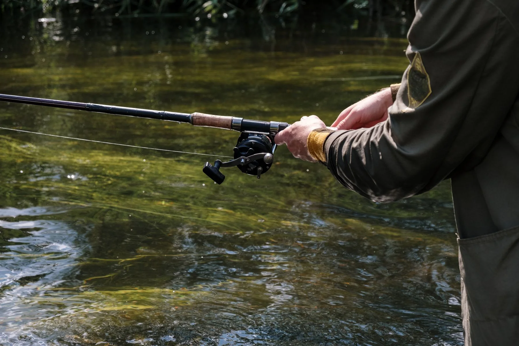 Person in a brown jacket holding a fishing rod and reel over water.